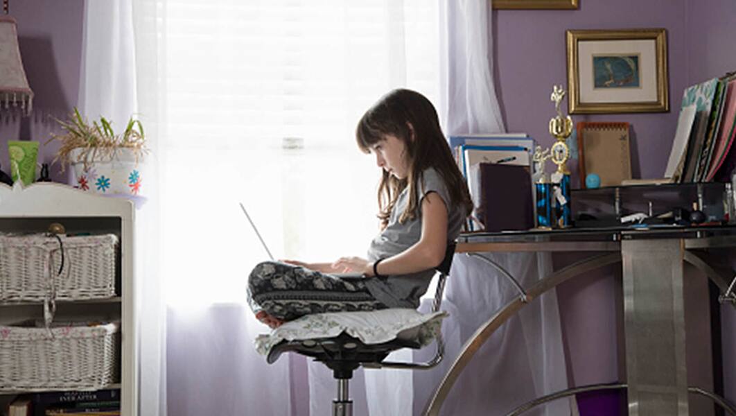 A young girl sits on a chair in a bedroom with a laptop on her lap
